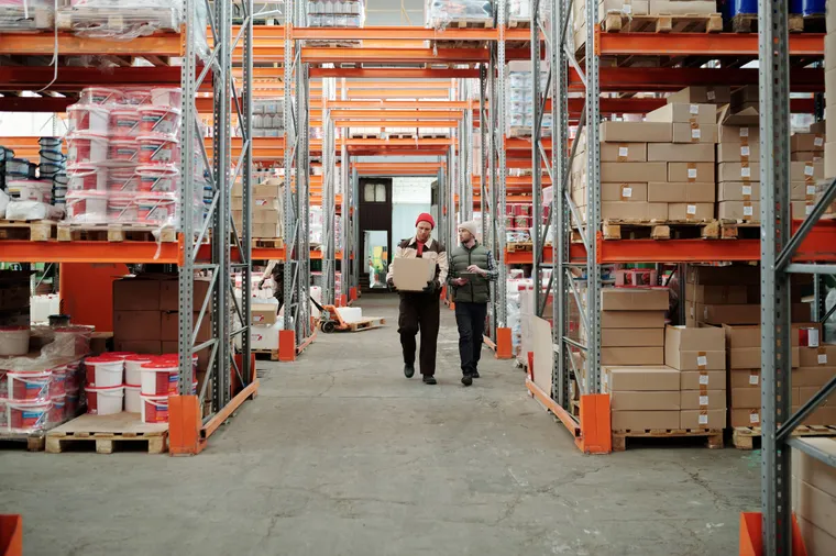 Dos trabajadores de almacén recorriendo un pasillo entre racks altos de mercancía paletizada